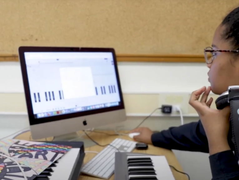 Student wearing headphones sits in front of music keyboard and monitor 