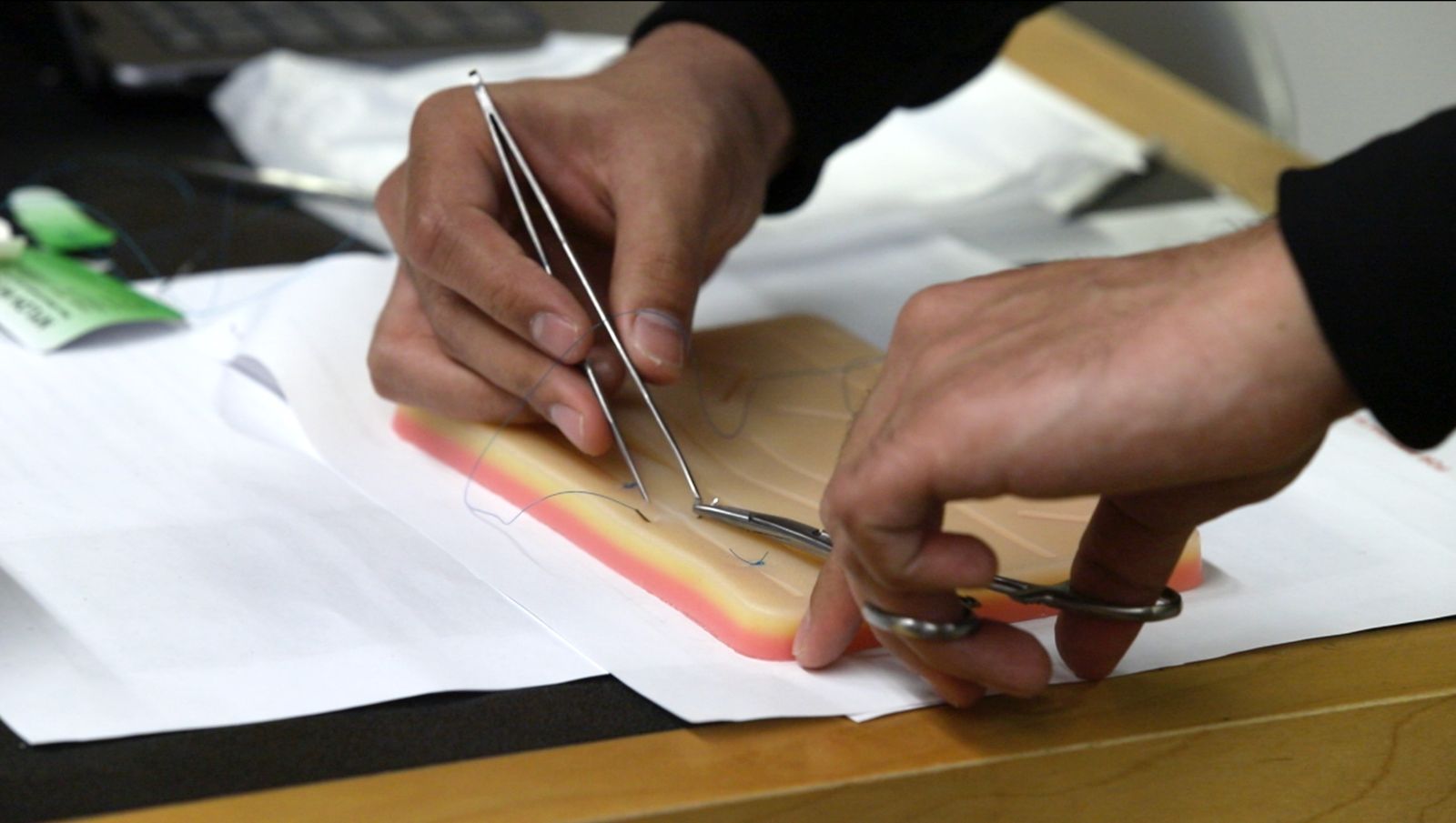 Hands shown using tools to surture on a mock object.