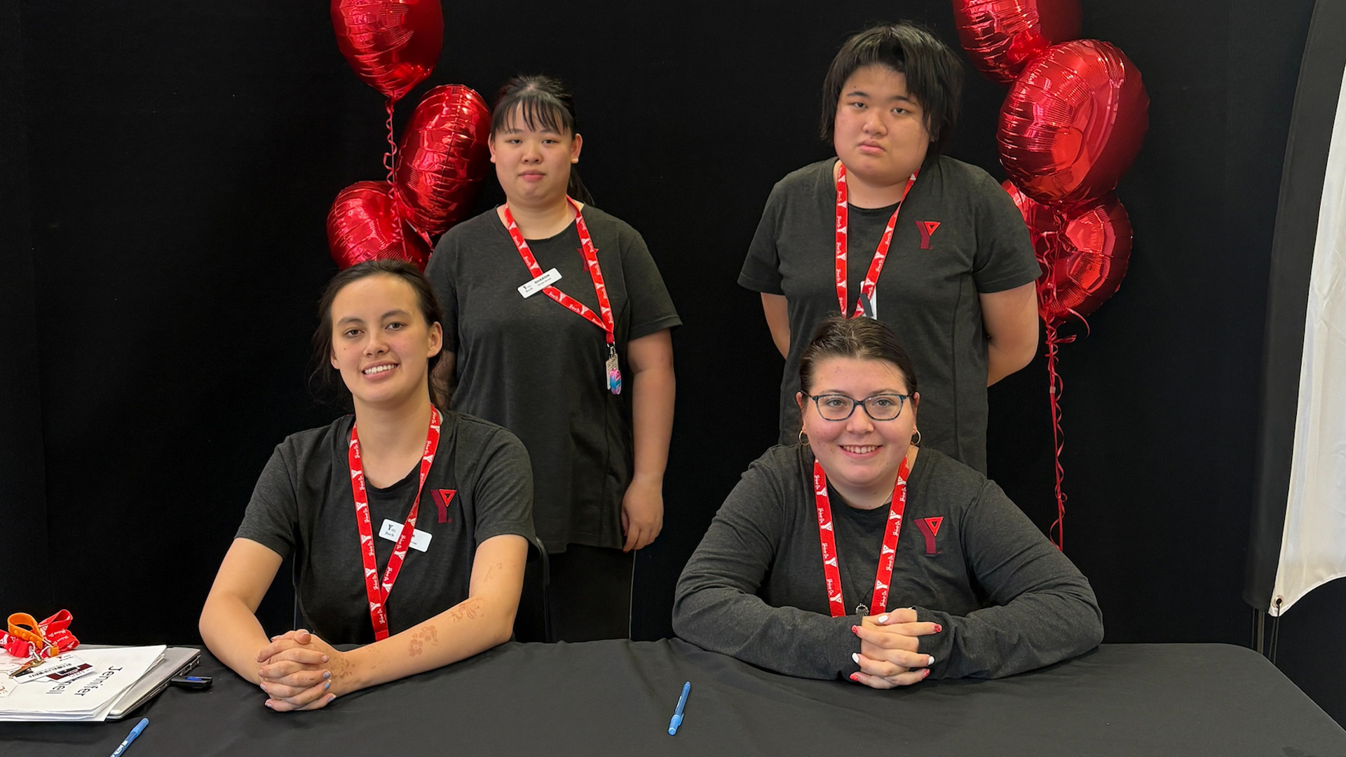 Two students sit, two stand behind them wearing black shirts and red lanyards. Sign behind them reads Let your potential shine.