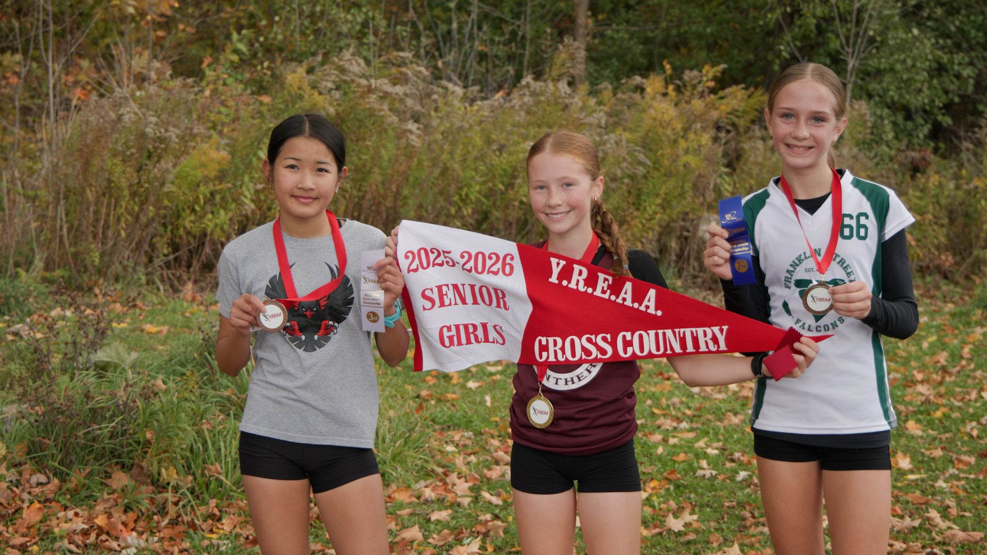 three students stand outside holding medals and a pennant that reads 2025-2026 Senior Girls YREAA Cross Country
