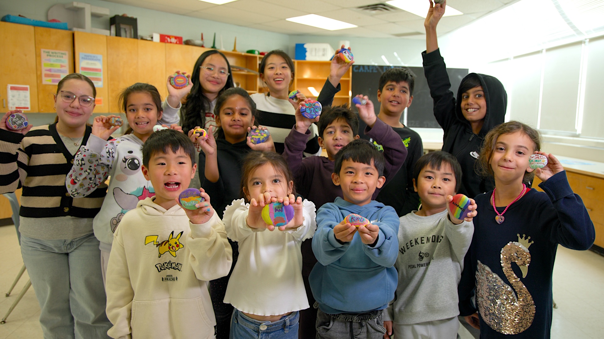 Students stand together in a group in a classroom holding up painted rocks