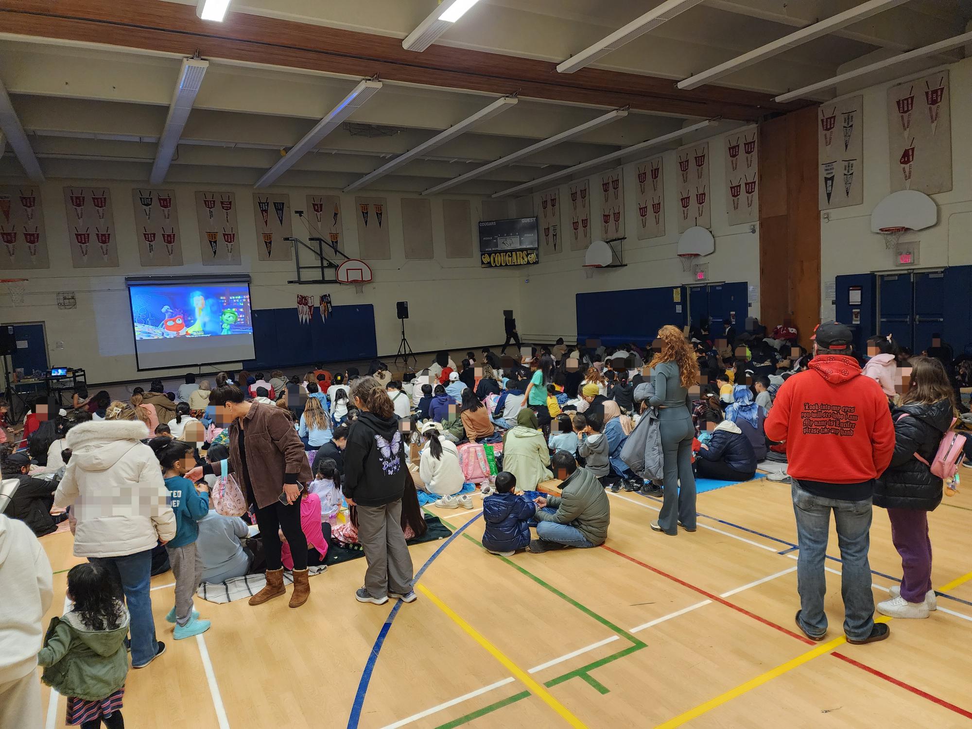 An image of families seated at the Crosby Heights Public School gym, participating in a movie night entertainment event, with a scene from the movie Inside Out showing on the screen projector.