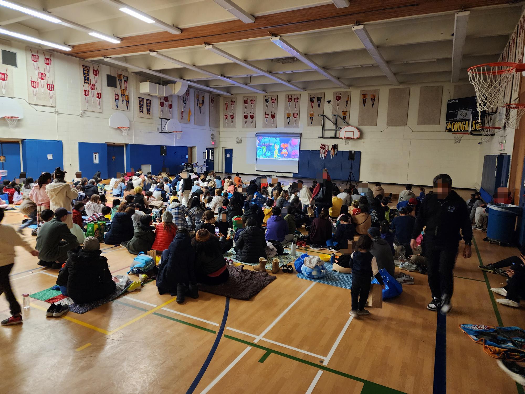 Another image of families seated at Crosby Heights Public School gym participating in a movie night entertainment event, with a scene from the movie Inside Out showing on the screen projector.