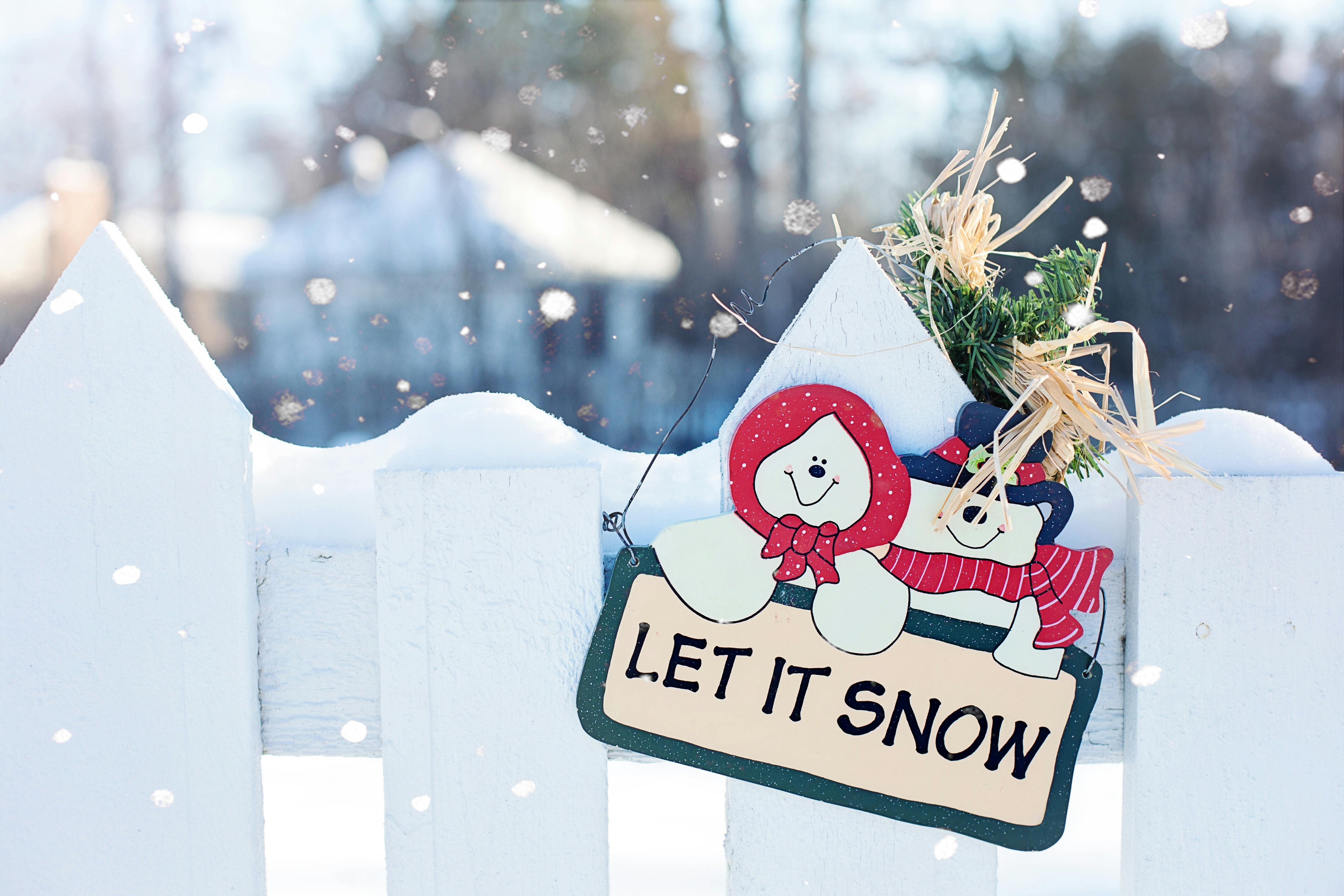 Image of a wintery scene outdoors with light snow falling on a white picket fence with a decorative ornamental sign that reads Let It Snow with a pair of decorative snowmen, one with a red bonnet and another with a red scarf and black hat..