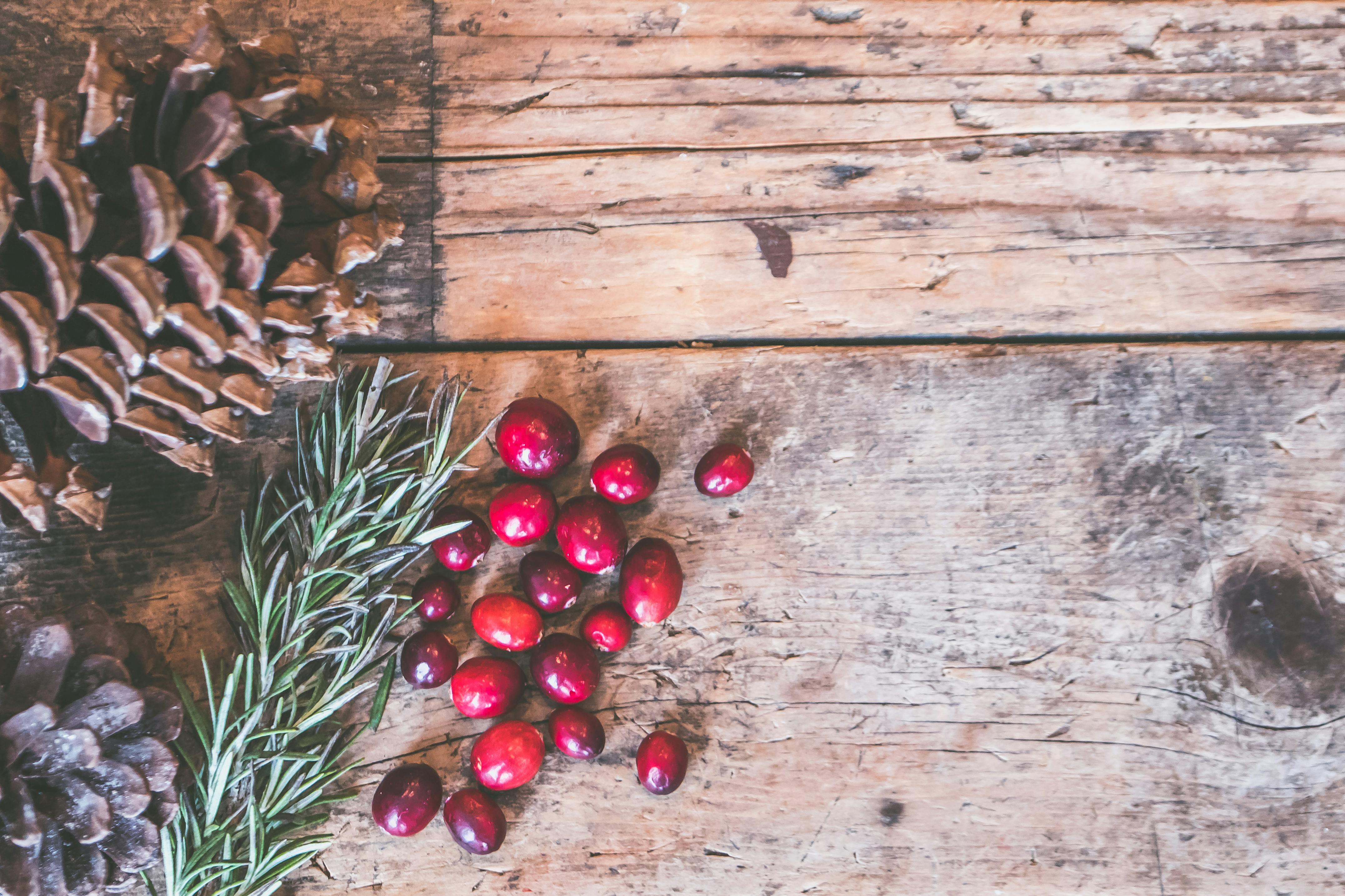 Image of a few pine cones, a sprig of rosemary, and a handful of red berries with a weathered wooden background to depict winter festive imagery