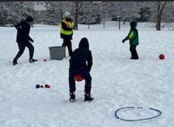 Wintery image of four individuals (two adults and two children) standing in a circle and playing a game with red balls using a grey tote as a makeshift ball basket outdoors in the snow
