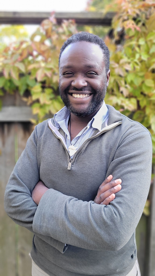 Image of Samuel Kwendo, a black man dressed in a grey sweater standing around trees with his  arms crosse in front on him
