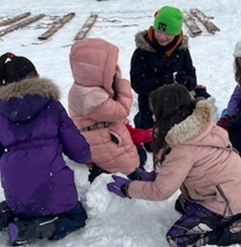 Image of four students dressed in their winter jackets with gloves kneeling on the snow building a snowman