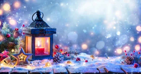 Winter scene of a picnic table covered with snow and decorated with a black lantern with a burning red candle surrounded by ornaments such as a lighted decorative star, bells, pine cones, berries, greenery.