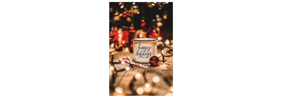 Holiday scene with a white melamine looking mug with black cursive script reading happy holidays sitting on a wooden picnic table with globe lighting and holiday ornaments decorating the table such as a candy cane, red bell-like christmas ornament and what looks like a blurred backdrop of a lighted christmas tree.