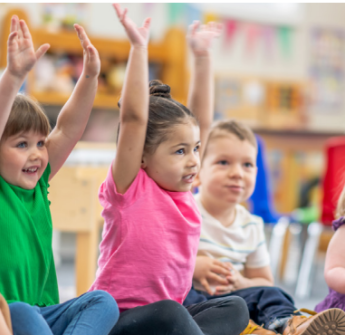 Elementary students sit in a classroom on the floor, some with their hands in the air