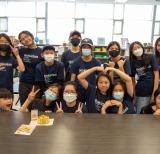Group of students wearing blue shirts stand in a group in front of a table in a classroom