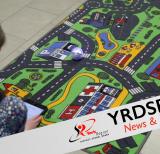 Student bent over floor mat featuring roadways, holding a tablet