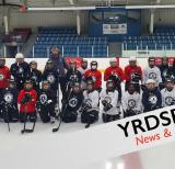 Students kneel and stand on the ice in a group holding hockey sticks wearing helmets and red and white jerseys. Several adults stand on the ice with them
