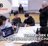 Students sit around table working on laptops with papers covering the table