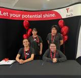 Two students sit, two stand behind them wearing black shirts and red lanyards. Sign behind them reads Let your potential shine.