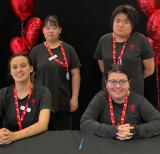 Two students sit, two stand behind them wearing black shirts and red lanyards. Sign behind them reads Let your potential shine.