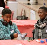 Parent and child sit at table covered in red table cloth. Child holds a ziploc bag with both hands. Measuring cups sit on the table. 