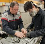 Student wears mask and works with supervisor on a car. They hold wires in their hands. 