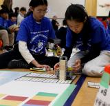 Two students wearing blue Tshirts kneel on the floor beside a small Lego robot on a mat. The students are focused on the robot. 