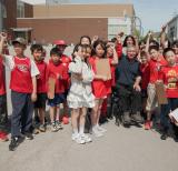 Students stand in a group around Rick Hansen outside the school, most are wearing red and lifting a hand in the air