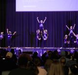 Students in cheerleading outfits stand in dance formation on a stage
