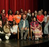 Elder and director sit in chairs. Students and adults stand and kneel around them on a stage