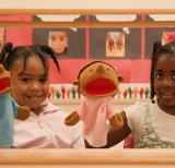Two students play with puppets between two red curtains of a puppet theatre