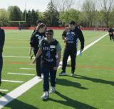 Four students stand on turf field smiling, walking towards the camera wearing blue sport shirts