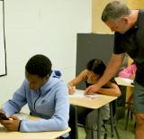 Teacher helps student at desk. Student in desk in front uses calculator.